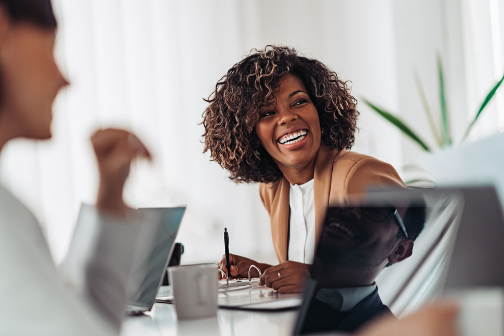 A beautiful woman, in a tan jacket, laughing in a meeting