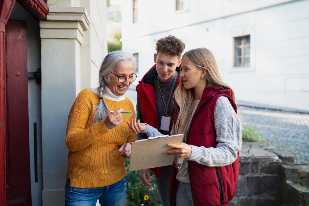 Young door to door volunteers talking to senior woman and taking survey at her front door.